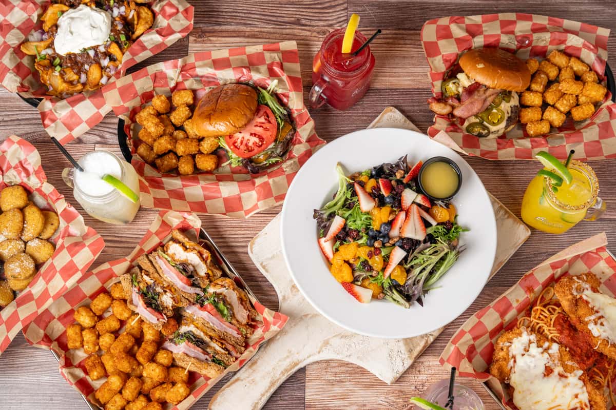 Overhead view of burgers, sandwiches, tater tots, salad, pasta, and colorful drinks on a wooden table.