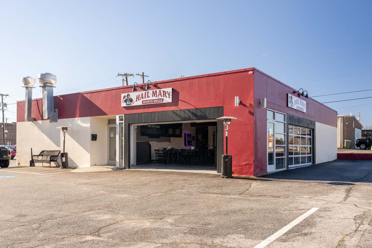 Exterior view of a red and white building labeled "Hail Mary Sports Grille," with large open garage-style doors, patio heaters outside, and a mostly empty parking lot under a clear sky.