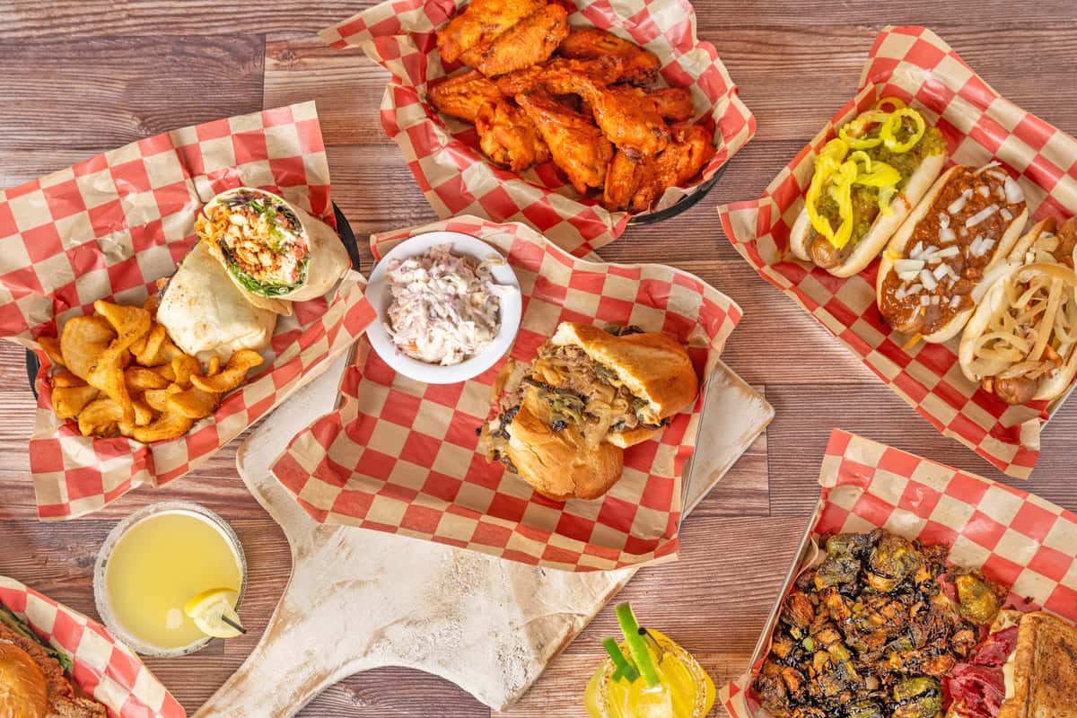 Overhead spread of wraps, cheesesteak, hot dogs, wings, fries, and coleslaw on red checkered paper.