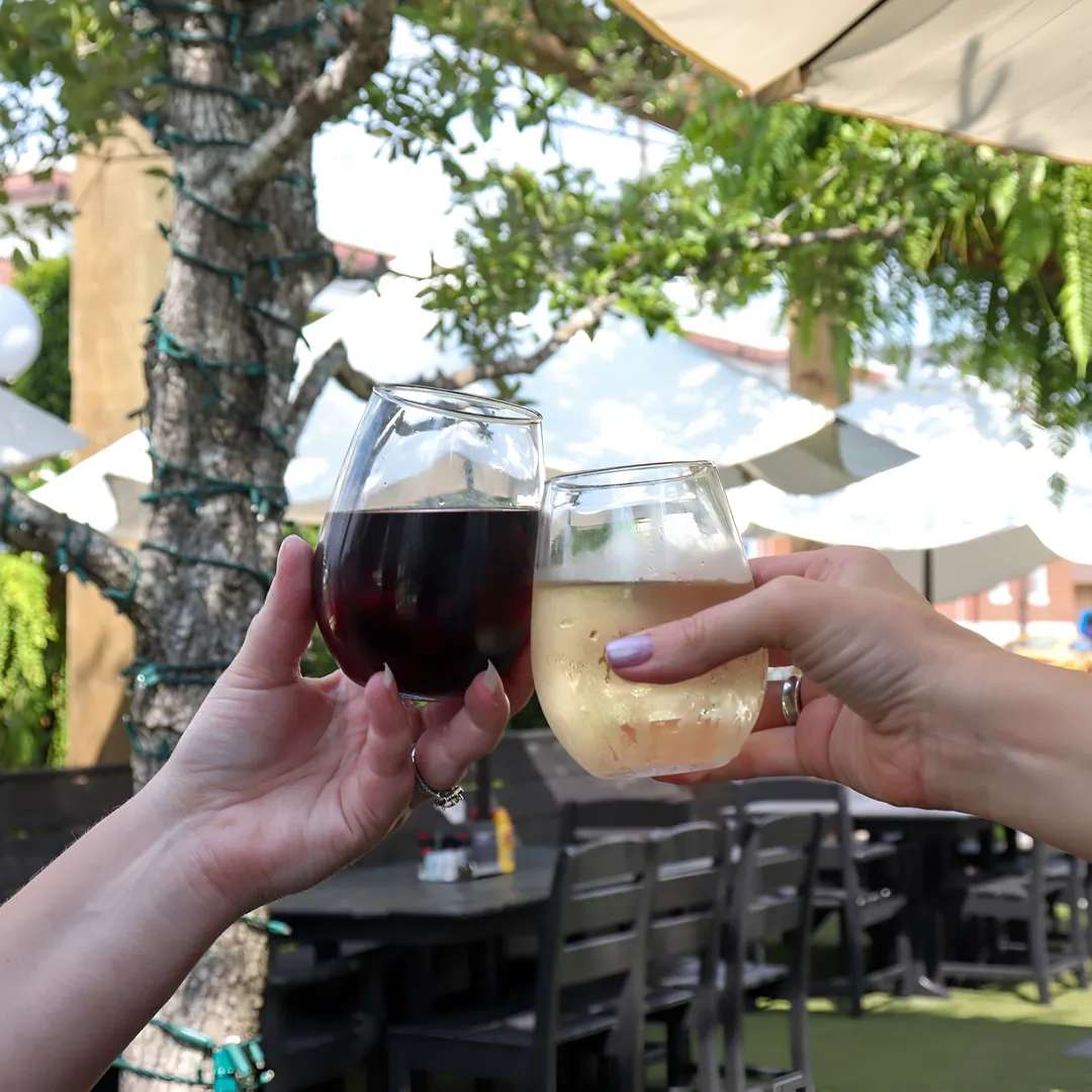Two people clinking glasses, one with red wine and the other with white wine, against a sunny outdoor background with patio seating.