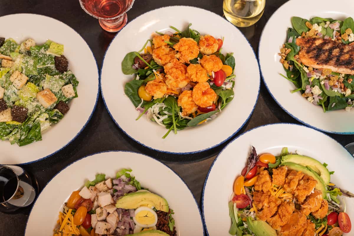 A variety of salads, including a Caesar salad, a shrimp salad with tomatoes, a grilled chicken salad with avocado, and a fried chicken salad, all served on white plates.