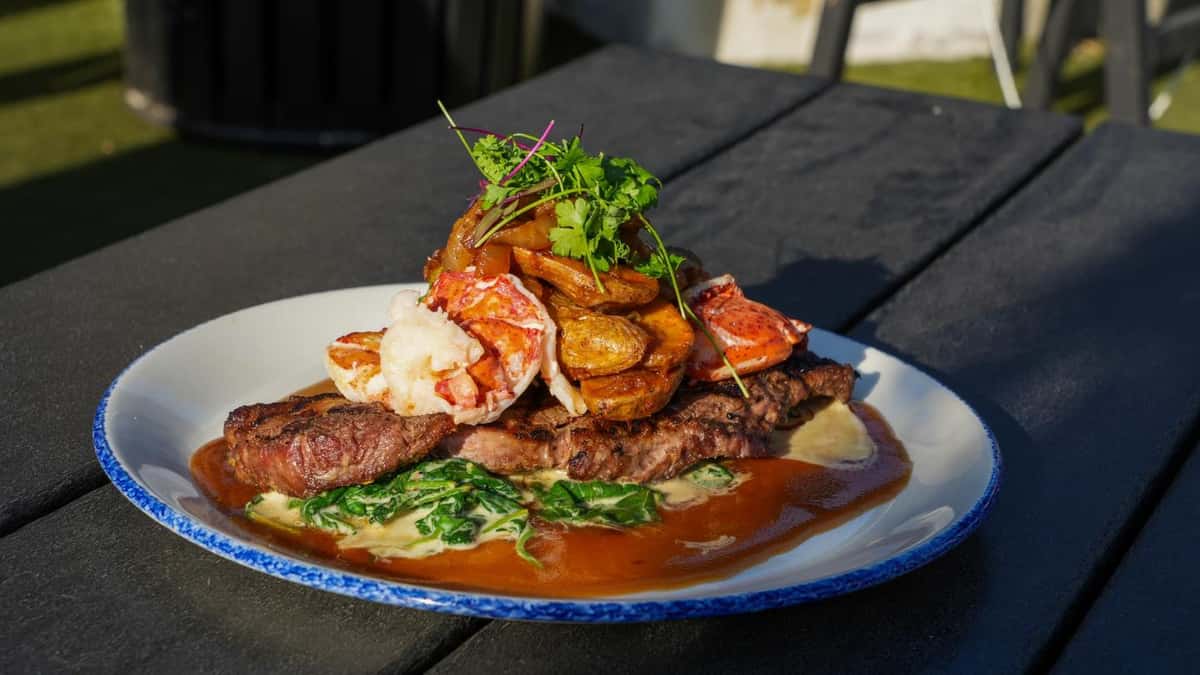 A grilled steak topped with lobster and crispy potatoes, served over a bed of creamy spinach and drizzled with a rich sauce. Garnished with fresh herbs.