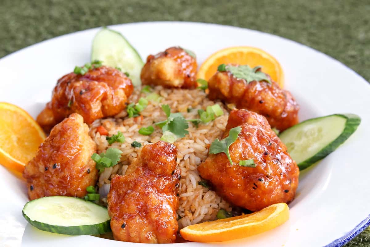 A plate of orange chicken served with seasoned rice, garnished with fresh cilantro, cucumber slices, and orange wedges.