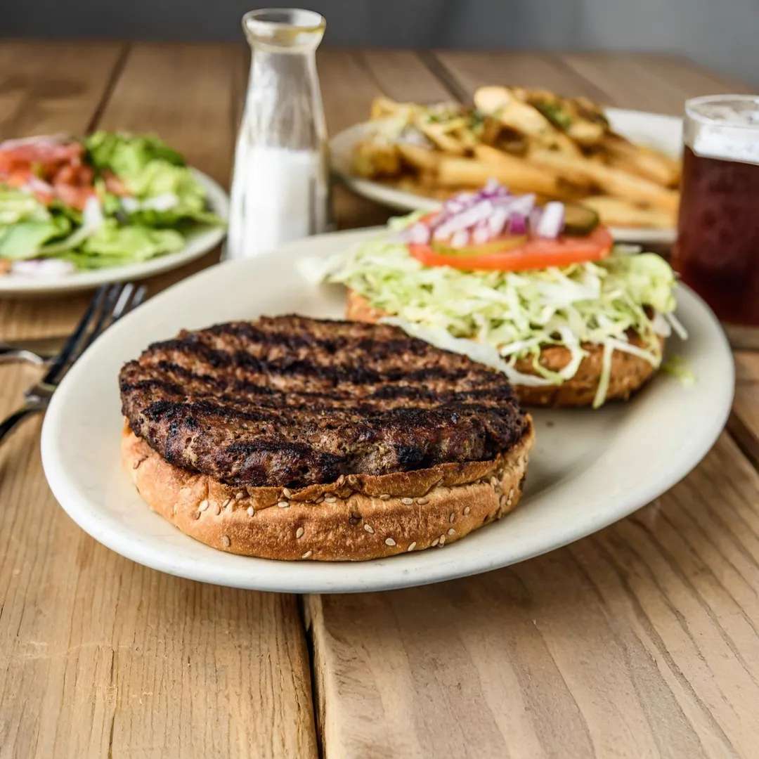 A grilled hamburger patty served on a sesame seed bun with lettuce, tomato, and onions, with a side of fries, salad, and a drink.