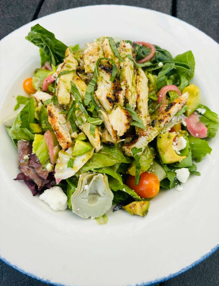 This appears to be the same grilled chicken salad shown previously — featuring sliced grilled chicken breast atop a vibrant mix of greens with ingredients like avocado, artichokes, cherry tomatoes, pickled onions, and crumbled feta, garnished with fresh basil strips. It looks both fresh and hearty! Let me know if you'd like help identifying ingredients, nutritional details, or recreating the dish.