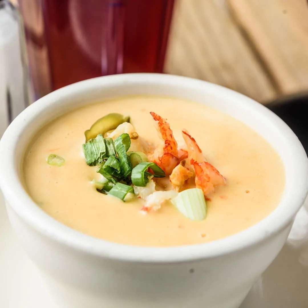 Creamy seafood bisque topped with green onions, lobster meat, and sliced pickles in a white bowl.