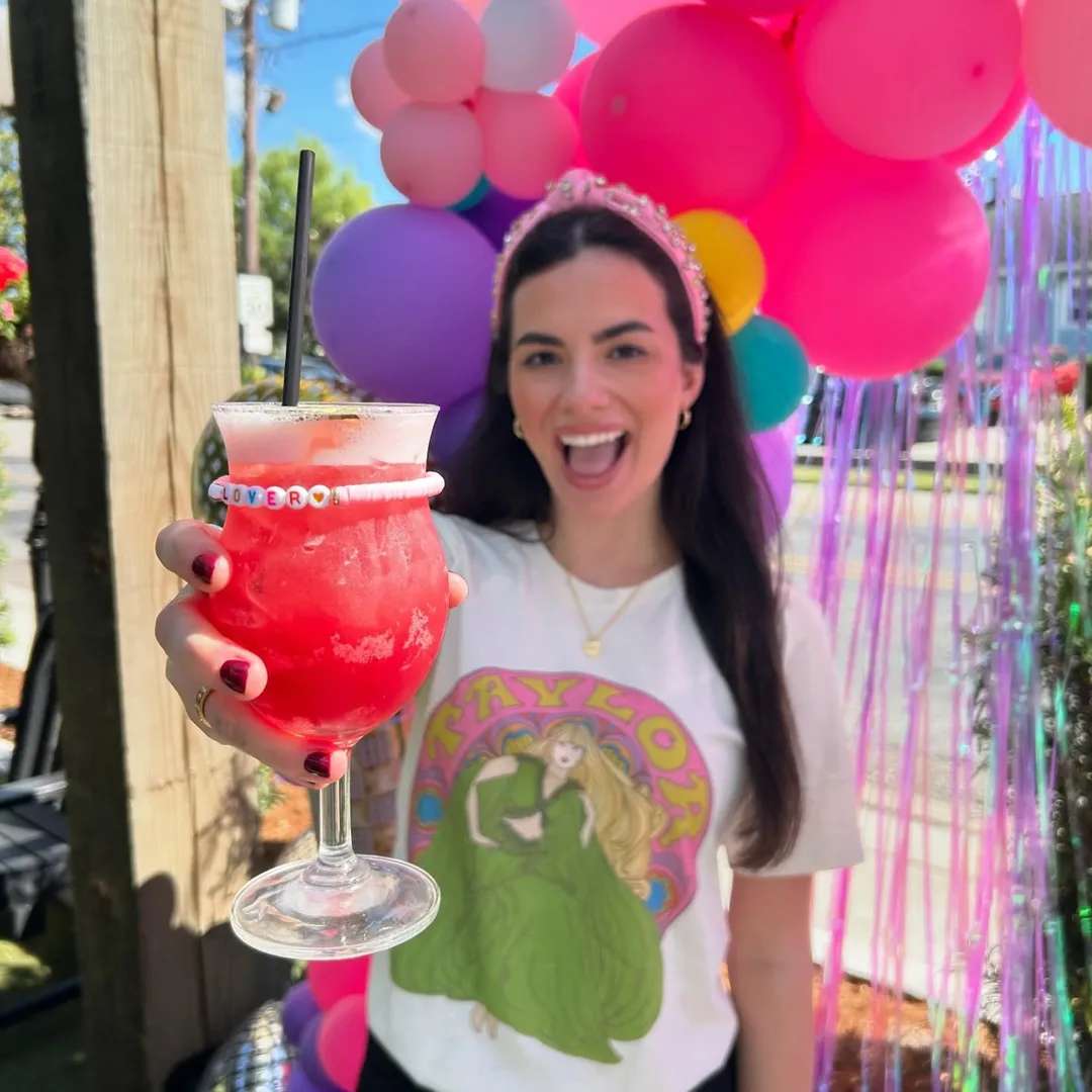 A woman holding a tropical cocktail in front of a vibrant balloon backdrop, wearing a "Taylor" t-shirt and a tiara, smiling excitedly.