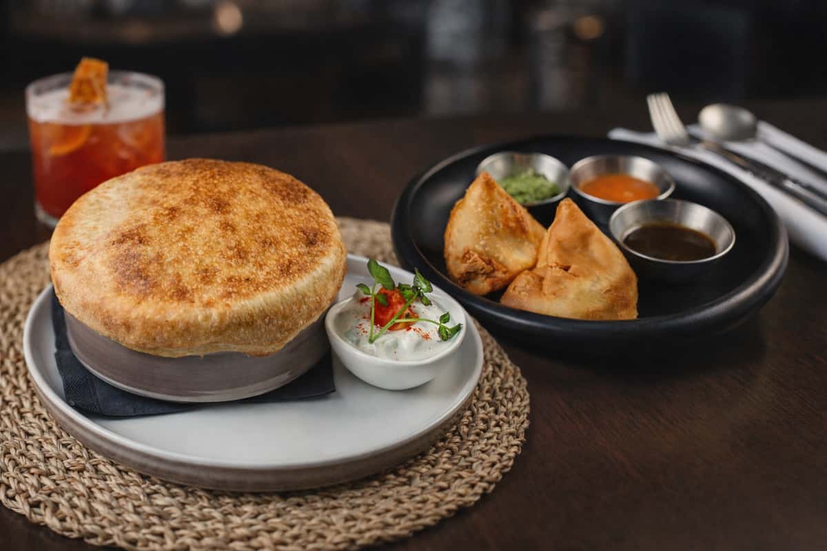 Golden baked savory pot pie with a flaky crust, served alongside crispy samosas and dipping sauces, highlighting modern Indian comfort food in a cozy upscale restaurant.