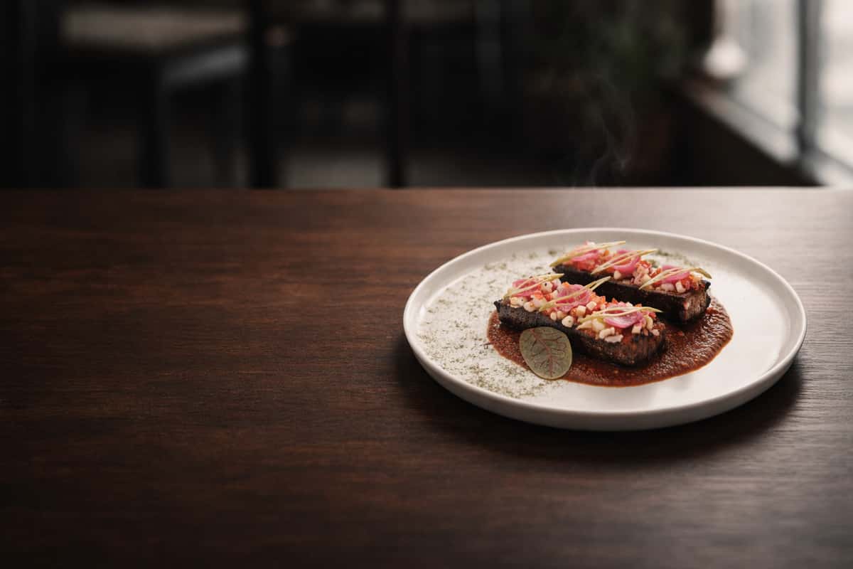 Seared beef or lamb bites topped with pickled onions and grains, plated over a rich brown sauce in a modern Indian fusion restaurant with warm ambient lighting.