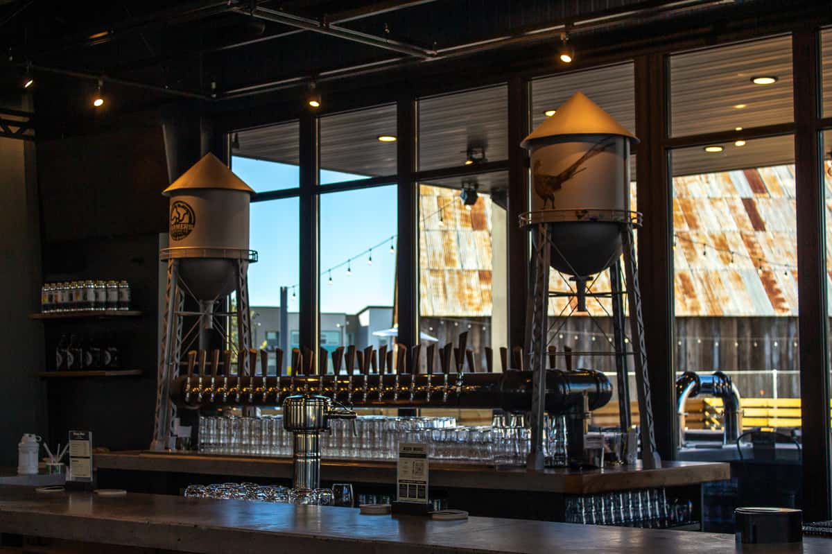 A modern taproom bar with rows of beer taps, glasses stacked below, and two silo-style tanks as decorative features.