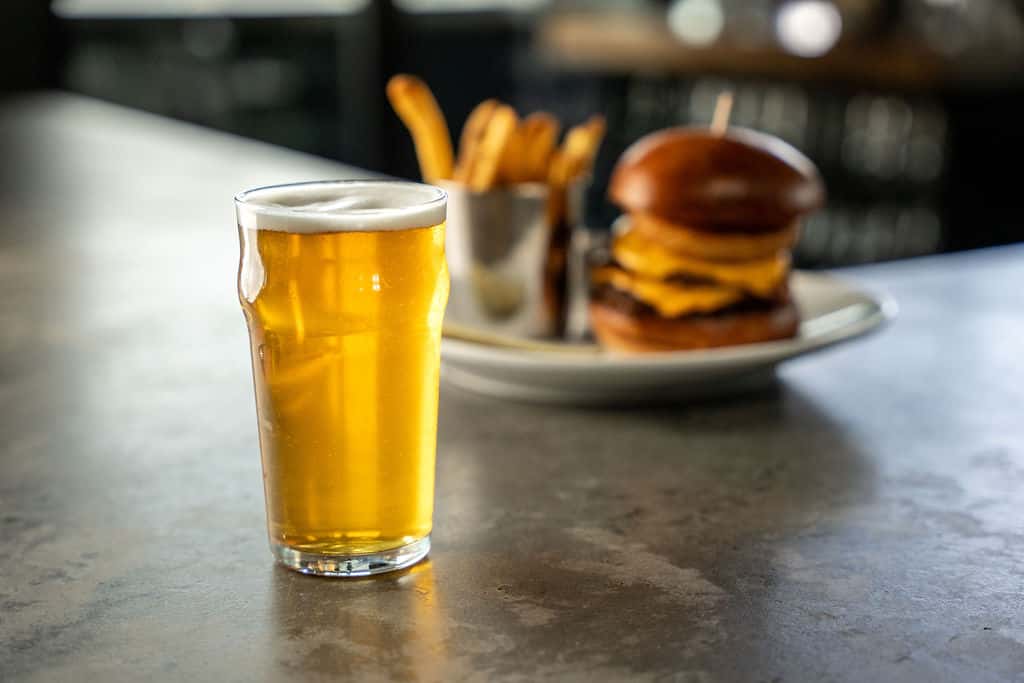 A pint of golden beer sits on a counter with a blurred burger and fries in the background on a white plate.