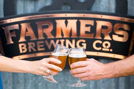 Two people clink beer glasses in a toast in front of a Farmers Brewing Co. sign.