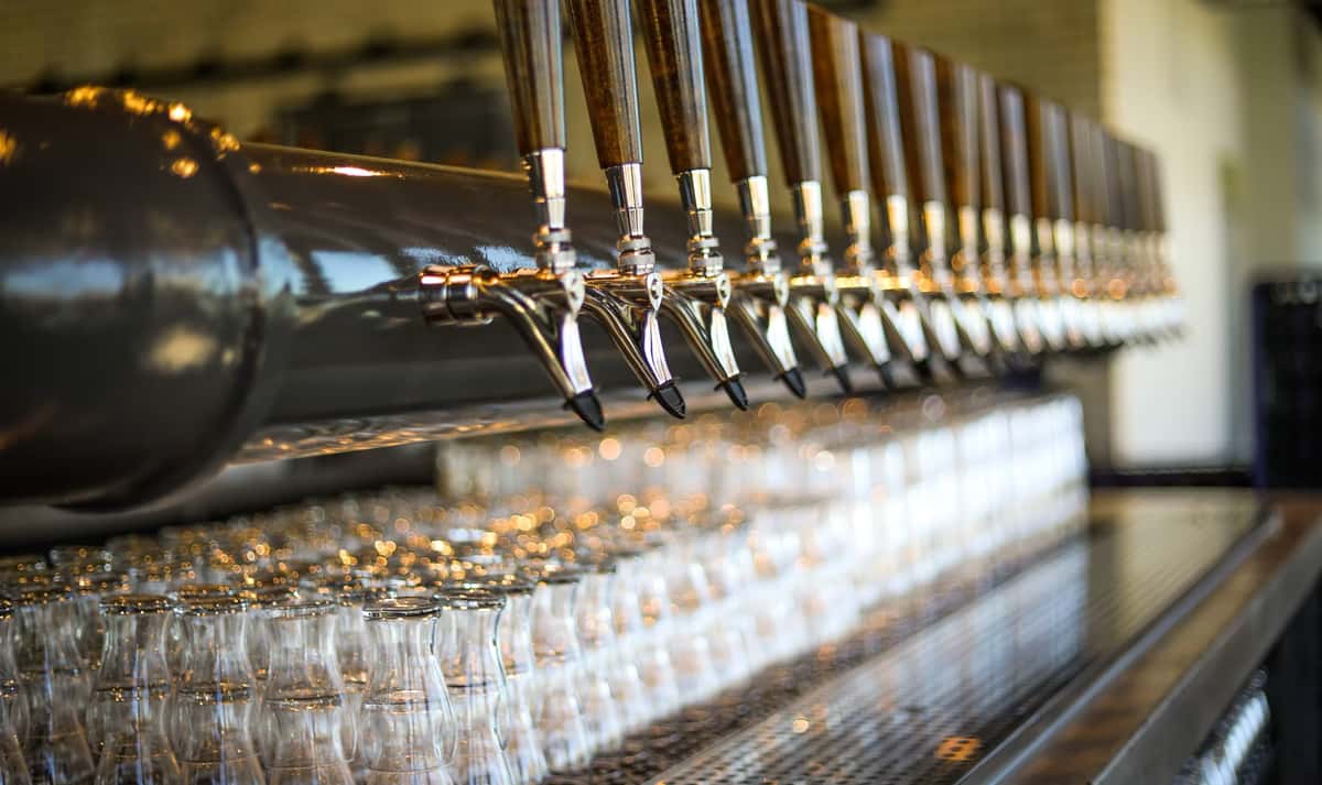 Row of shiny beer taps with wooden handles above neatly stacked glassware at a brewery bar.