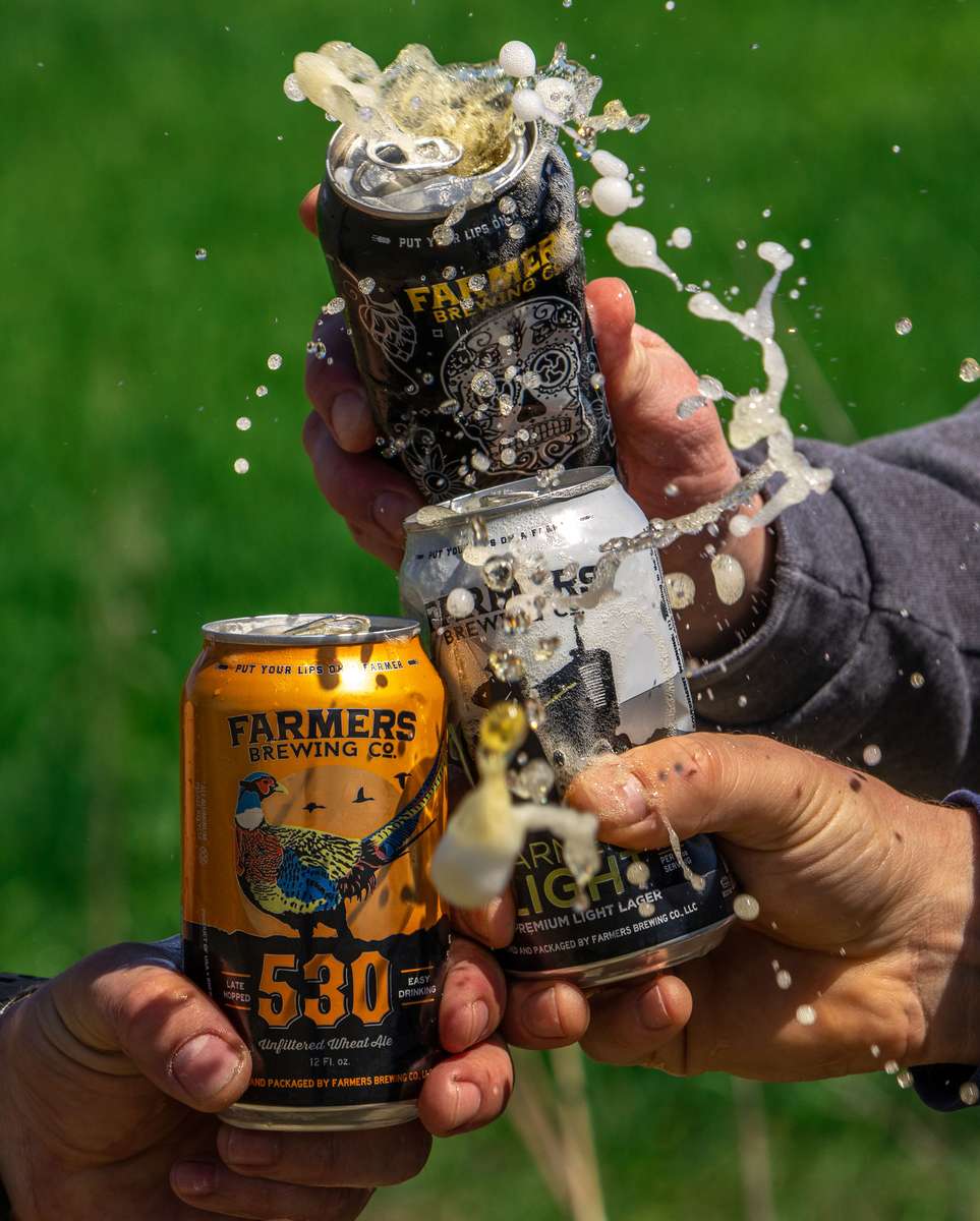 Hands clinking Farmers Brewing Co. beer cans with foam and beer dramatically spraying into the air.