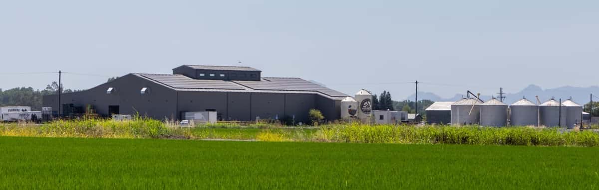 A large brewery building with silos sits beside green fields under a clear sky with distant mountains.