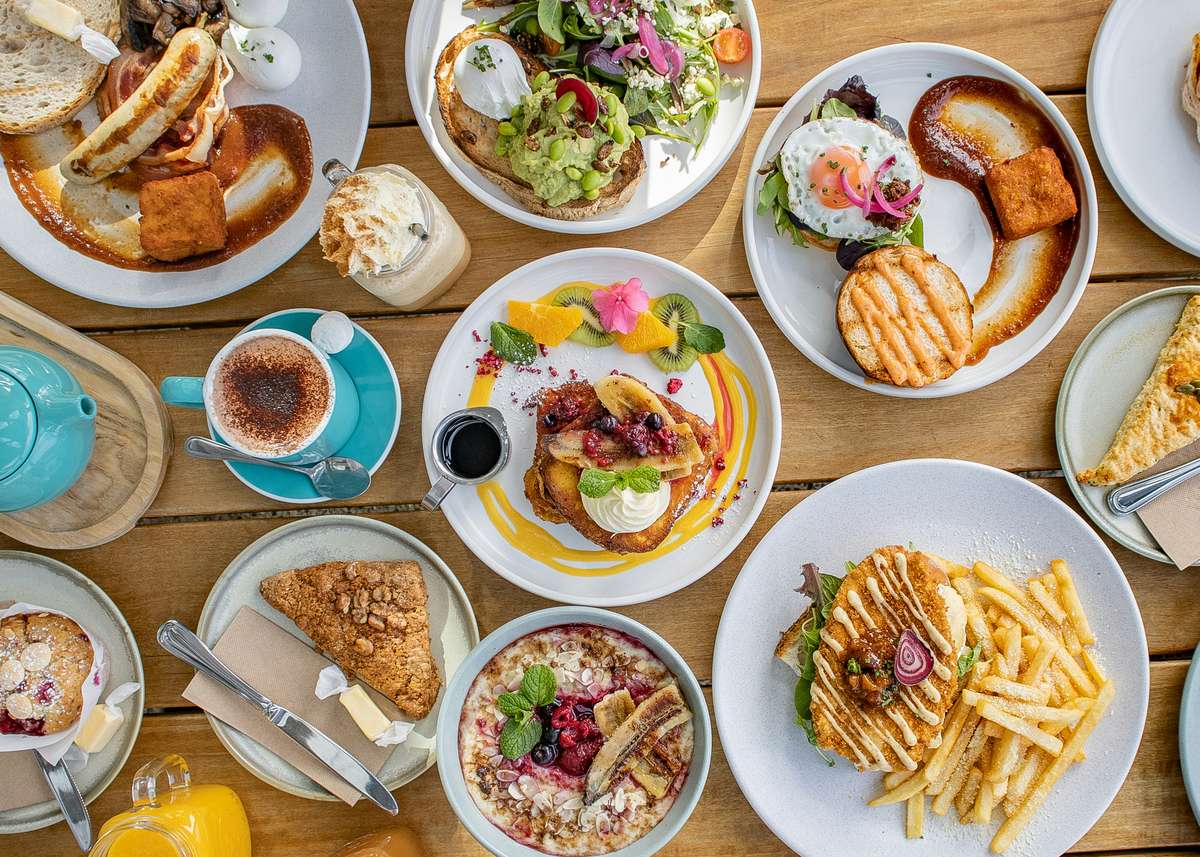 Colorful brunch spread with pancakes, toast, fries, salads, coffee, juice, and desserts on a wooden table with vibrant plating.