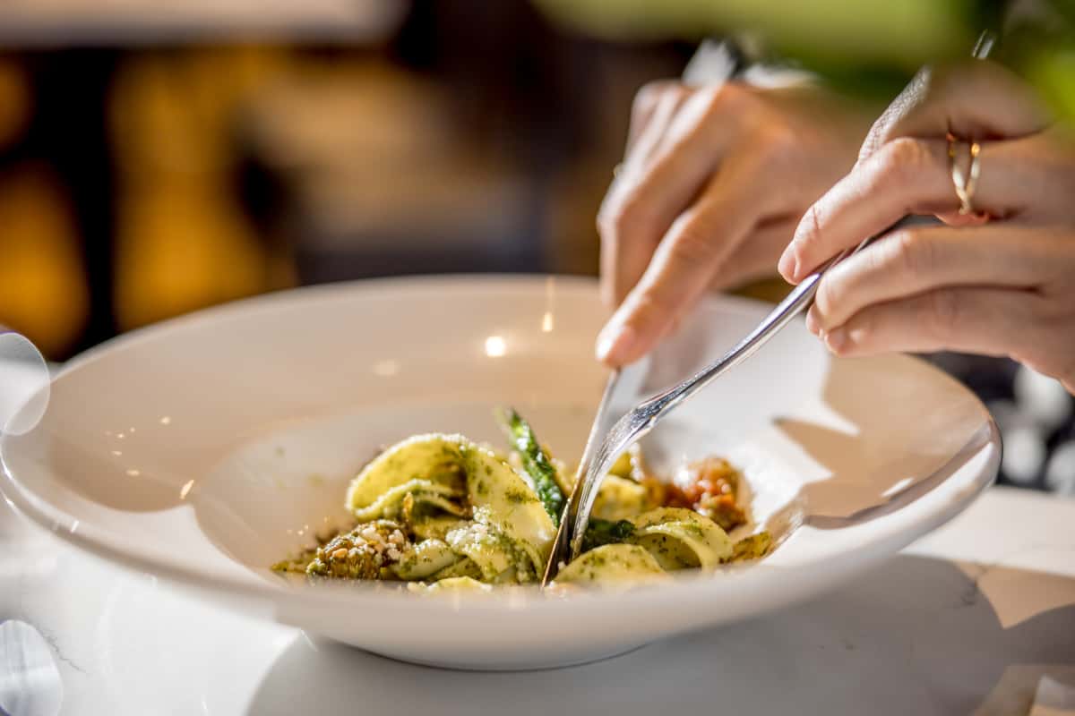 Person using fork and knife to eat pasta with green pesto sauce and vegetables in a white bowl on a white table.