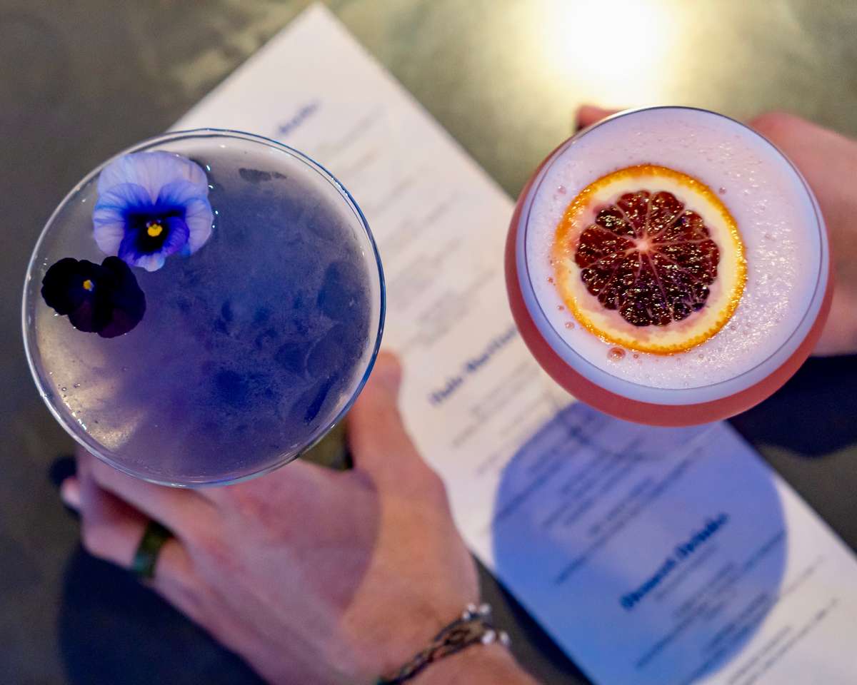 Close-up of two colorful cocktails, one with edible flowers and the other topped with a blood orange slice, held above a drink menu.