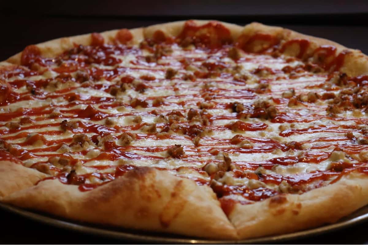 This is a close-up image of a pizza with a golden, fluffy crust. The toppings include a layer of melted cheese, scattered pieces of sausage or ground meat, and a drizzle of red sauce (possibly BBQ or ketchup) over the top. The pizza appears to be freshly baked and served on a round metal tray.