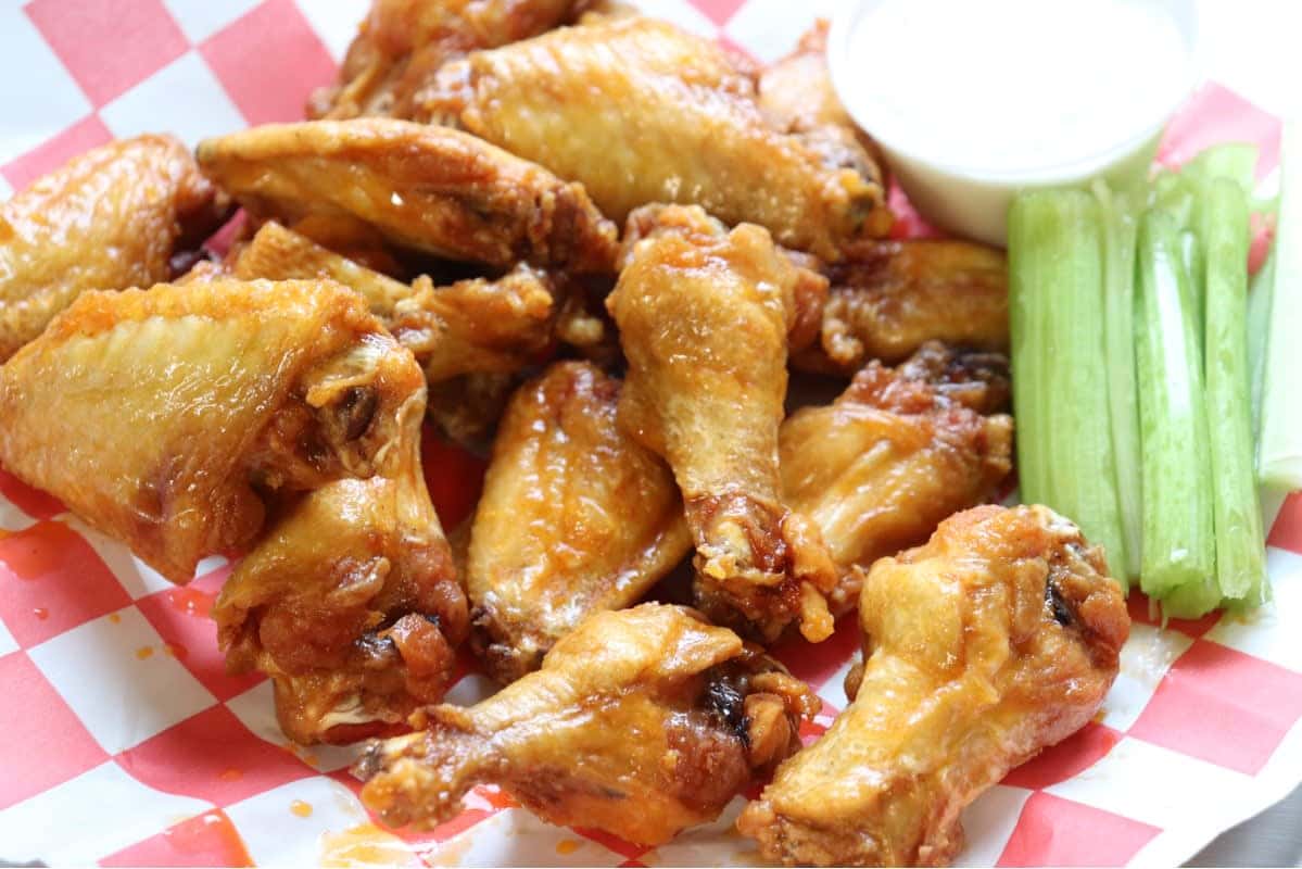 Golden fried chicken wings with celery sticks and a cup of ranch dressing on red-and-white checkered paper.