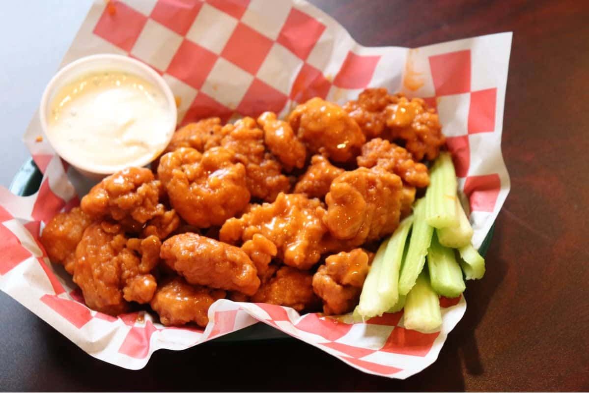 Buffalo boneless wings served with celery sticks and ranch dressing on red-and-white checkered paper in a green basket.