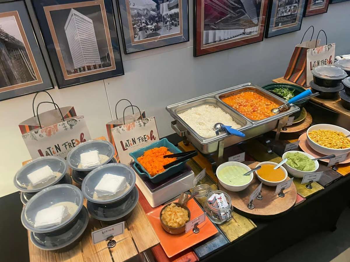 Latin buffet catering spread with white rice, beans, fresh toppings, shredded carrots, corn salsa, and house-made sauces, arranged on a colorful rustic display table.