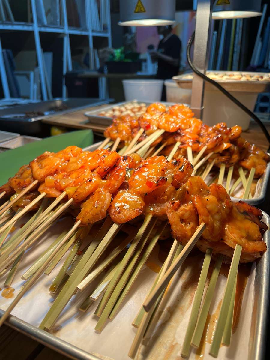 Grilled shrimp skewers glazed in a rich, spicy sauce, served hot on a catering tray under warming lights in a restaurant kitchen setting.