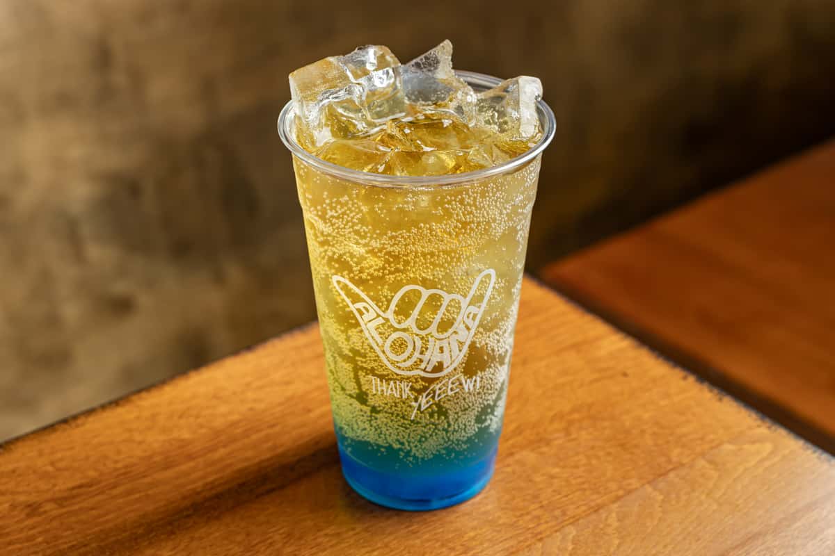 This is a sparkling drink served over ice in a branded ALOHANA cup. The drink features a vibrant gradient from blue at the bottom to golden yellow at the top, creating a refreshing tropical appearance.