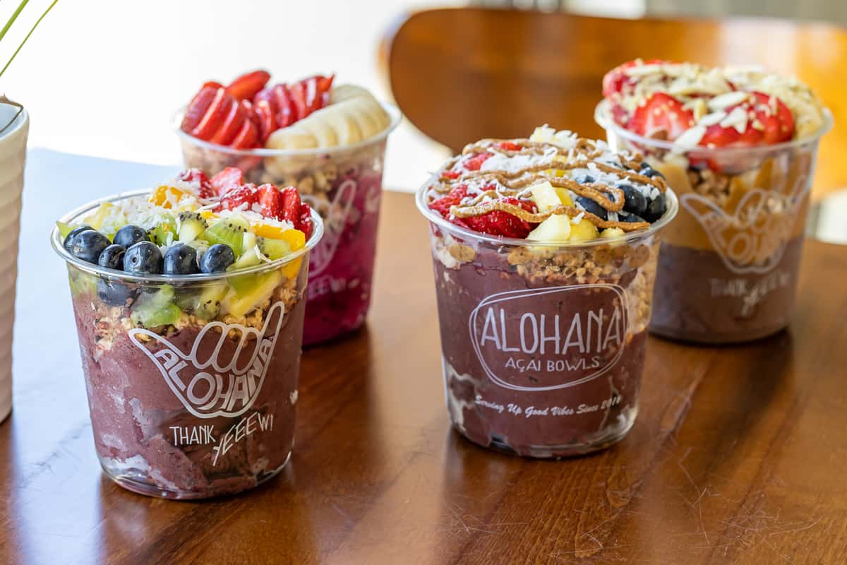 Four colorful acai bowls topped with fresh fruit, granola, and coconut flakes, arranged in a square on a wooden table.