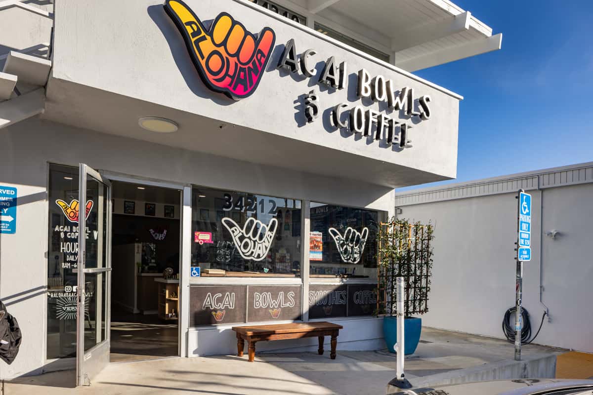 Exterior view of ALOHANA Açaí Bowls & Coffee shop with open glass doors, hand sign logos, and bold signage promoting açaí bowls, smoothies, and coffee, set against a bright blue sky in a coastal town.