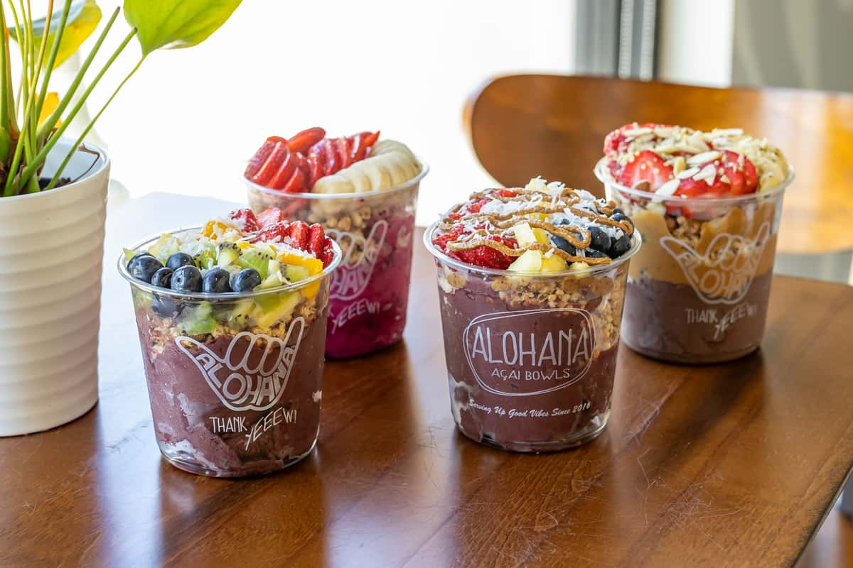 Four colorful acai bowls topped with fresh fruit, granola, and coconut flakes, arranged in a square on a wooden table.