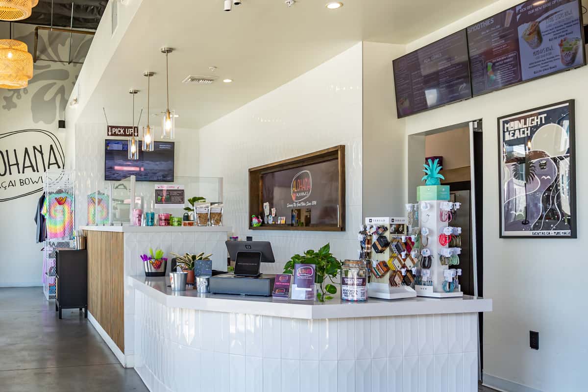 ALOHAHA counter with a register, colorful cups, keychains, plants, and a "Serving San Elijo Since 2020" sign in the background.