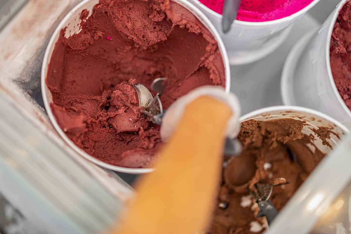 A gloved hand scoops dark red acai sorbet from a container in a freezer, surrounded by other vibrant sorbet flavors like pitaya (pink) and chocolate.