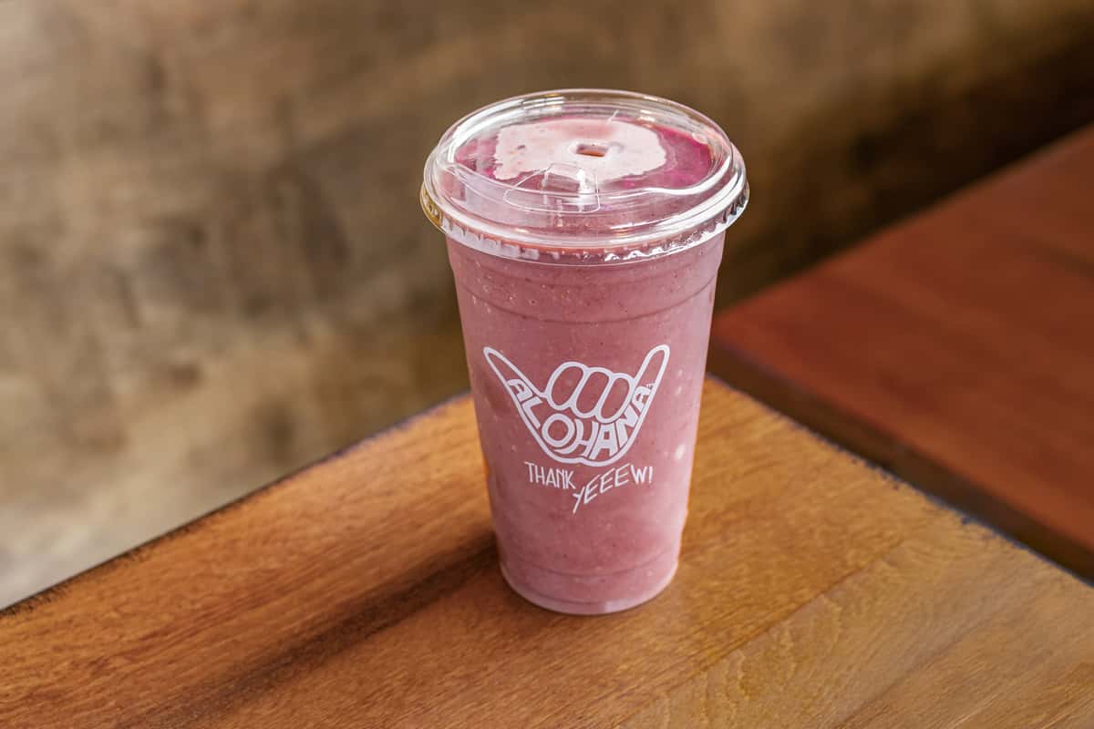 A thick, vibrant purple smoothie—likely made with berries or acai—is served in a clear cup branded with the "Alohaha" logo, sitting on a wooden table.