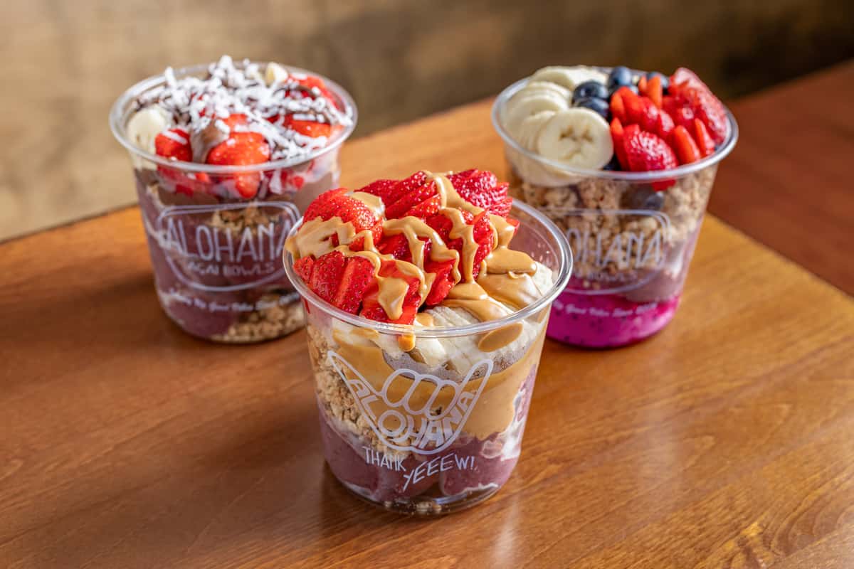 Three acai bowls topped with banana, strawberries, blueberries, peanut butter, and shredded coconut on a wooden table.
