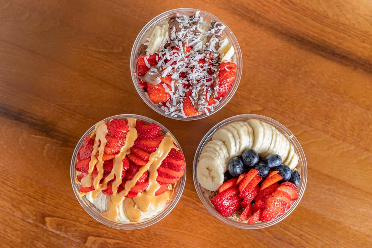 Three acai bowls topped with banana, strawberries, blueberries, peanut butter, and shredded coconut on a wooden table.