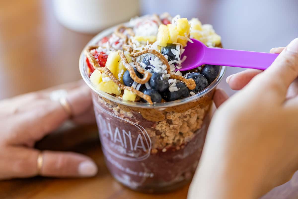 Close-up of a colorful acai bowl topped with pineapple chunks, blueberries, strawberries, shredded coconut, and granola, being scooped with a bright purple spoon.