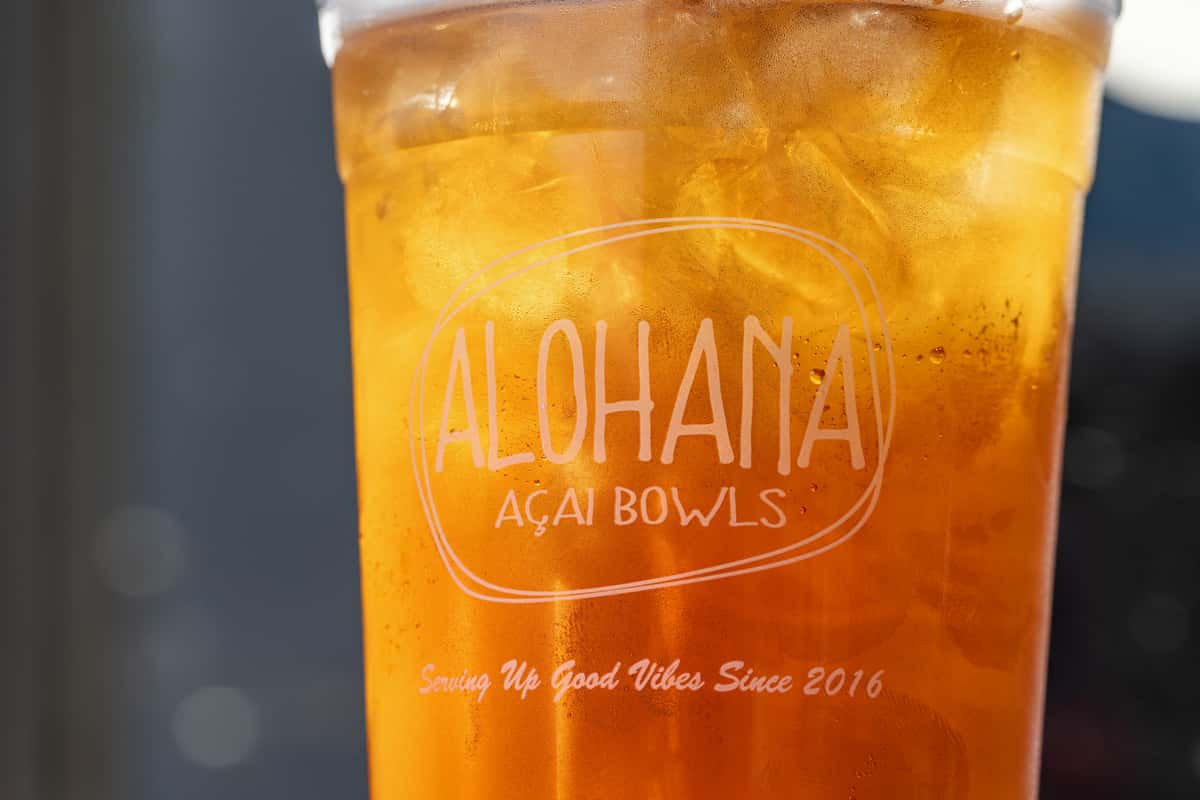 Close-up of a refreshing iced tea or tropical drink in a clear ALOHANA Açaí Bowls cup, filled with golden ice and condensation, highlighting the brand's "Serving Up Good Vibes Since 2016" slogan.