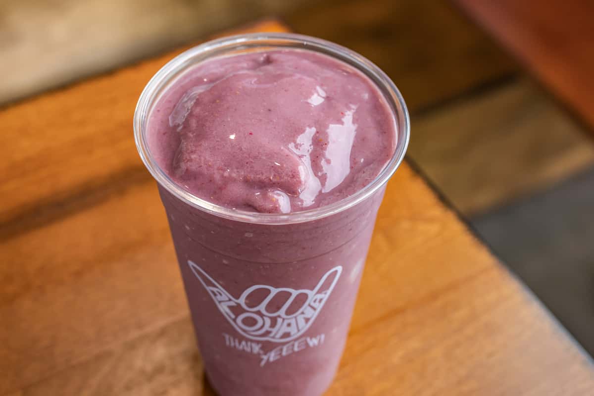 A thick, vibrant purple smoothie—likely made with berries or acai—is served in a clear cup branded with the "Alohaha" logo, sitting on a wooden table.