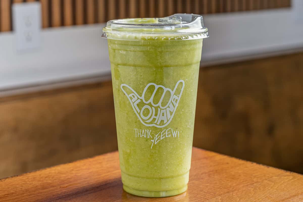 Green smoothie in a clear ALOHANA cup with lid, sitting on a wooden table against a wood-paneled wall.