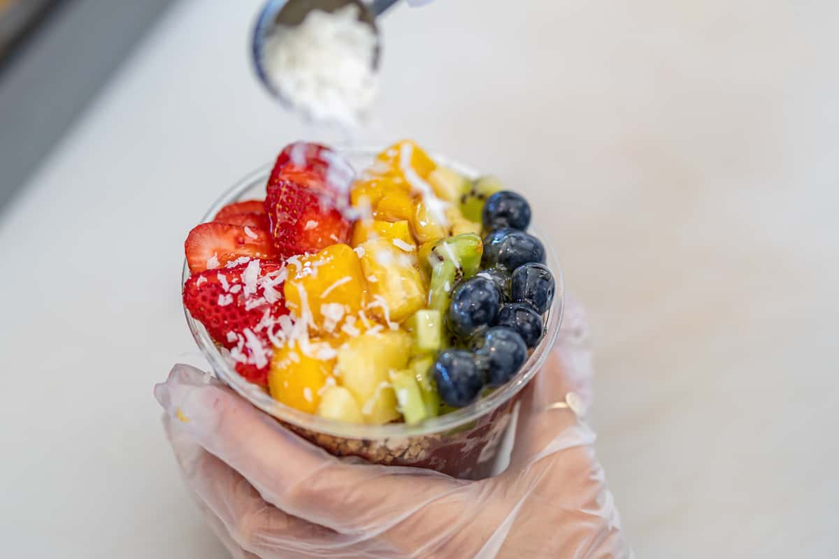 Gloved hand holds an acai bowl as shredded coconut is sprinkled over strawberries, mango, kiwi, pineapple, and blueberries.