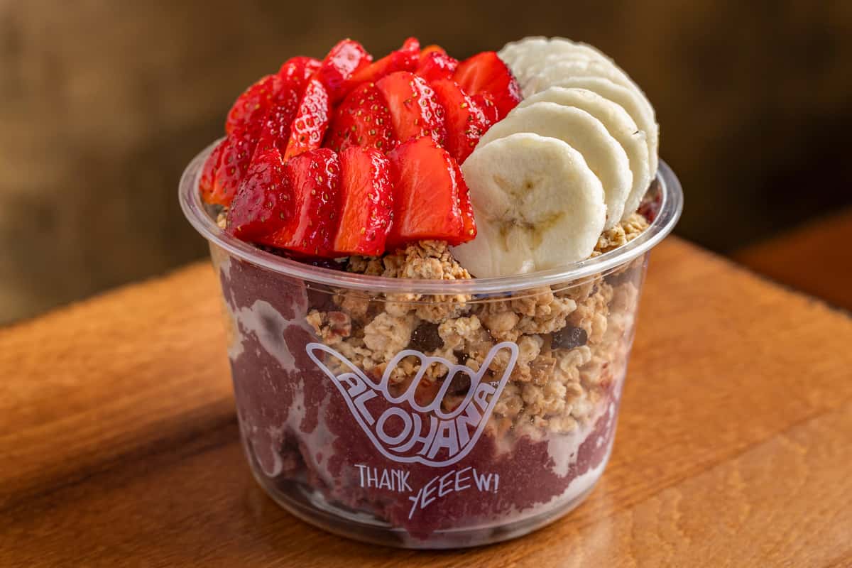 Bright pink smoothie bowl topped with sliced strawberries, bananas, and granola in a clear cup with "Alohana" branding.