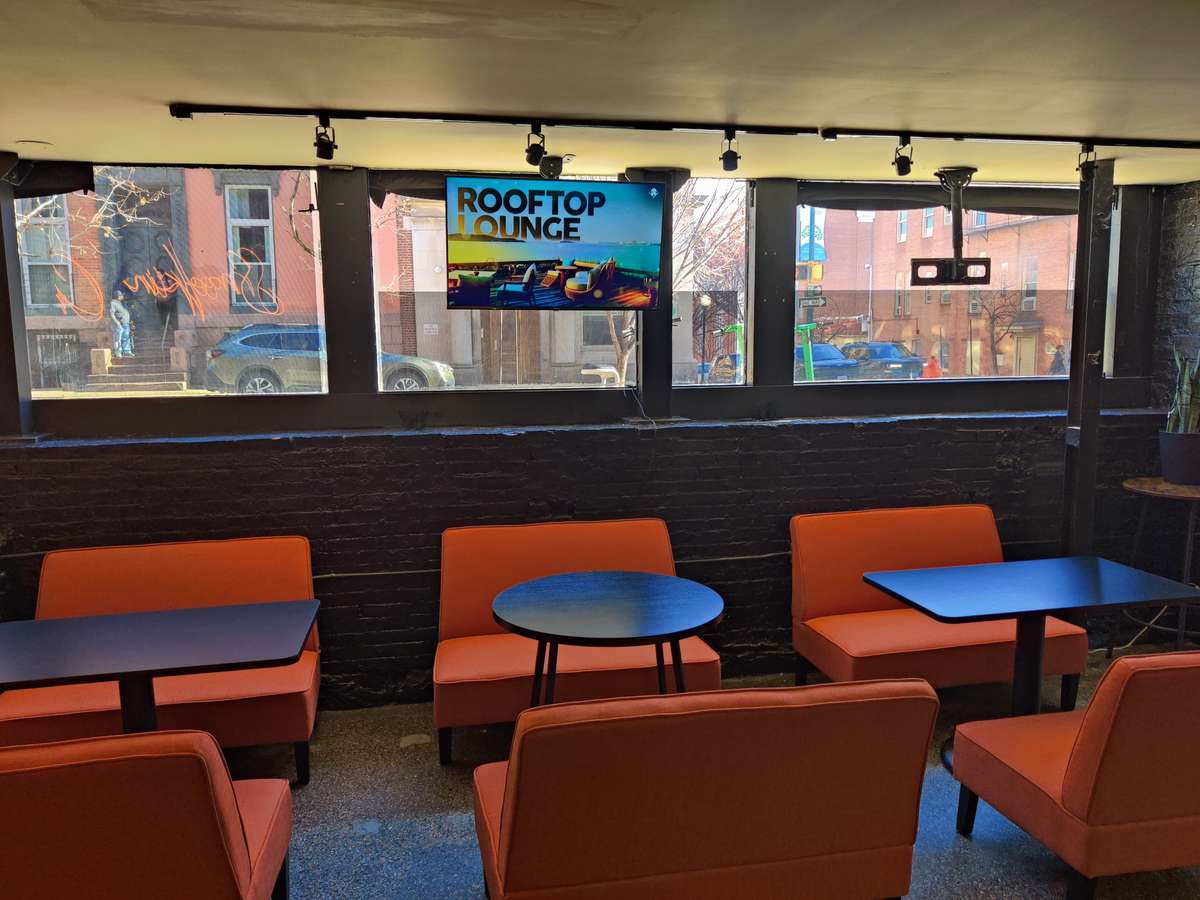 Modern casual restaurant lounge with orange chairs, small café tables, black brick walls, and large street-facing windows, with a TV display promoting a rooftop lounge.