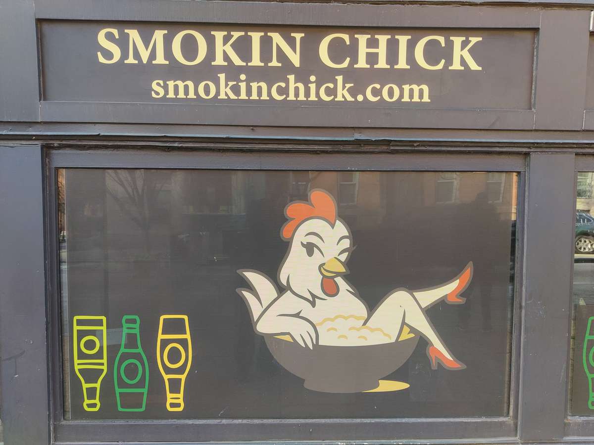 Restaurant storefront window with **"Smokin Chick"** signage and website URL, featuring a playful cartoon chicken lounging in a bowl graphic alongside bottle icons, branding for a casual BBQ or chicken restaurant.