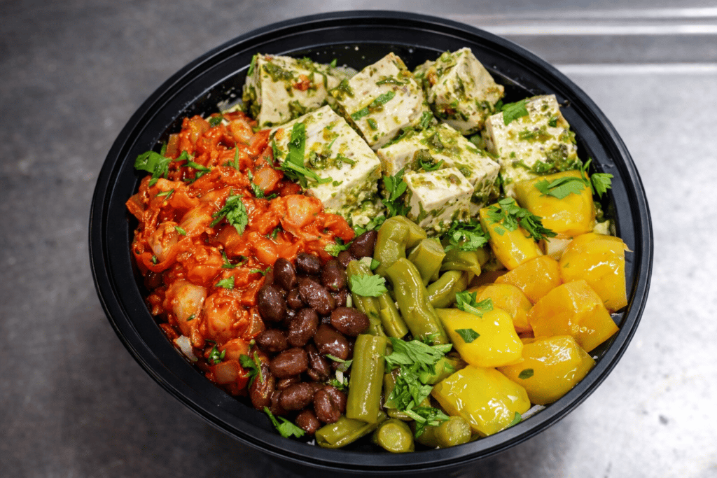 Colorful vegetarian grain bowl with herb-marinated tofu, kimchi, black beans, pickled vegetables, and fresh herbs over rice in a modern fast-casual restaurant bowl.
