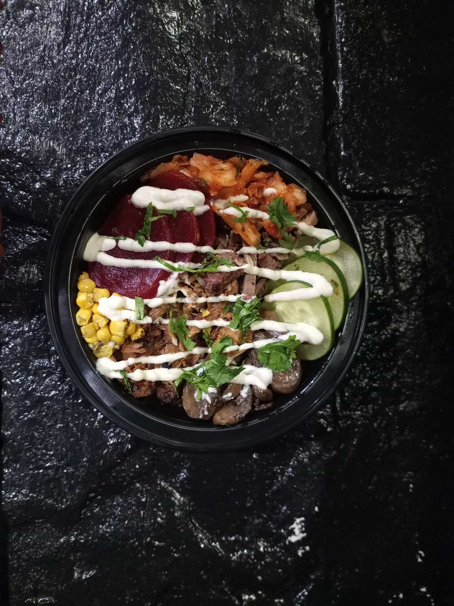 BBQ rice bowl with smoked pulled pork, roasted corn, sautéed mushrooms, cucumber slices, pickled beets, kimchi, and fresh herbs, finished with a creamy sauce drizzle.