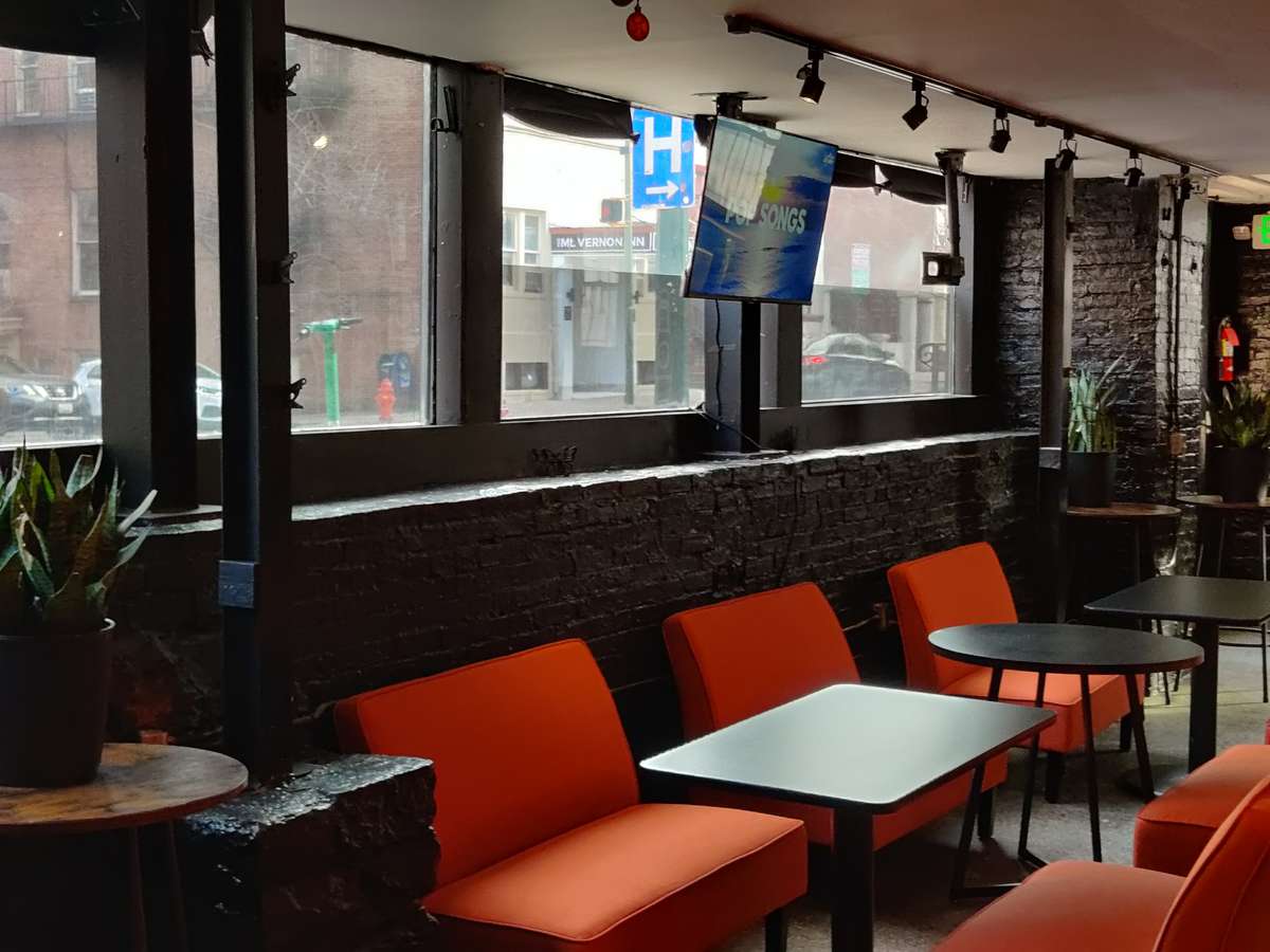 Modern casual restaurant interior with large street-facing windows, black brick walls, orange lounge chairs, and small café tables creating a relaxed urban dining atmosphere.
