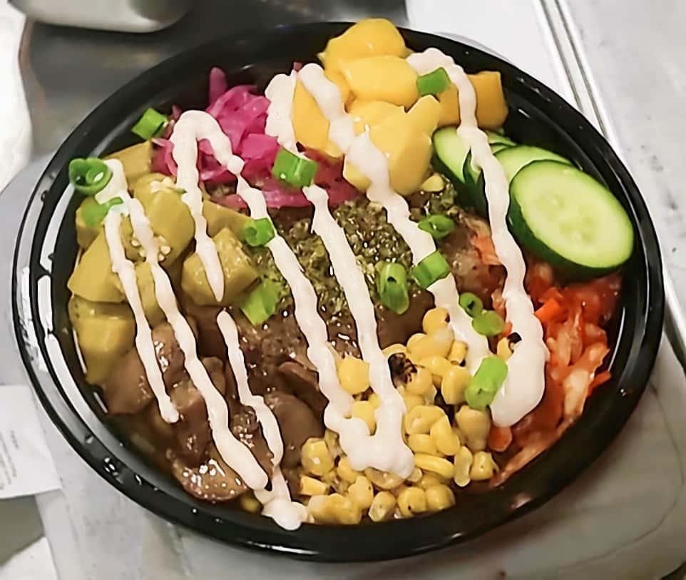 Grain bowl with grilled steak, avocado chunks, pickled red onions, mango salsa, roasted corn, cucumber slices, and scallions, drizzled with creamy sauce in a colorful modern restaurant bowl.