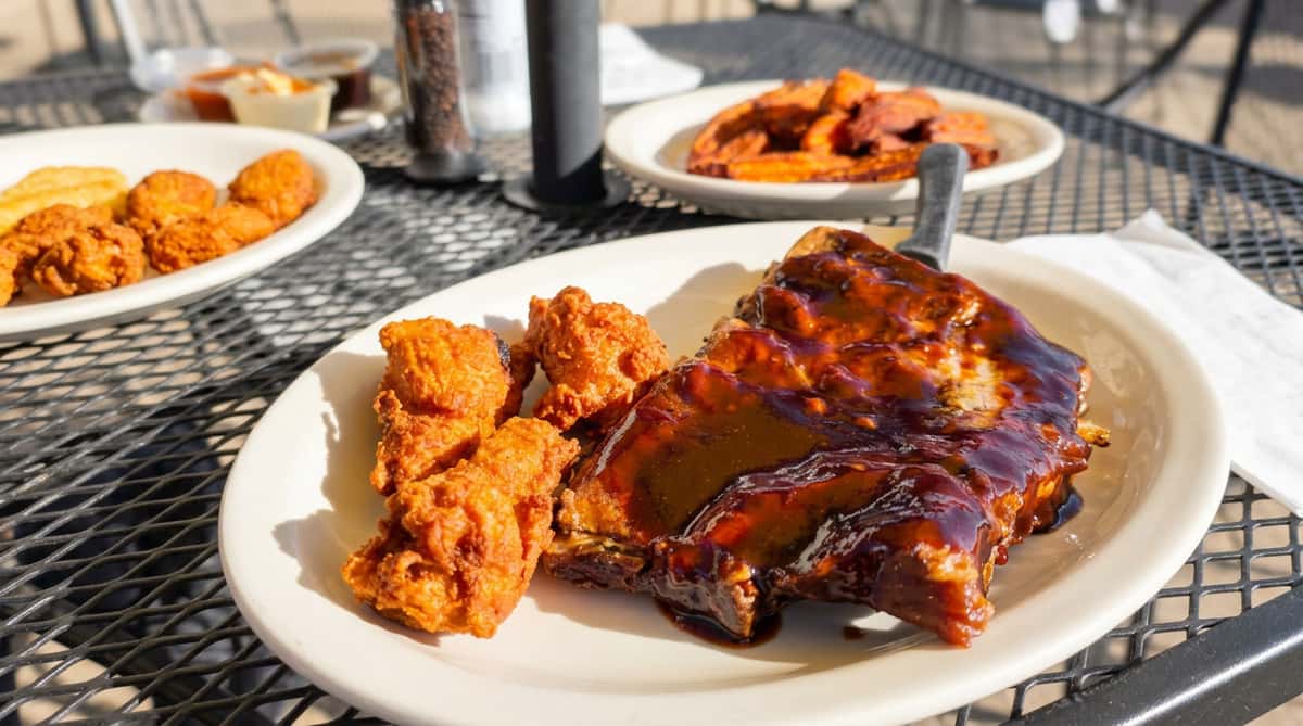 Ribs with BBQ sauce and fried chicken pieces on a white plate, with sweet potato fries in the background.