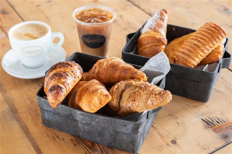 bread and pastries on a wood table with a cup of hot coffee and a cup of iced coffee.